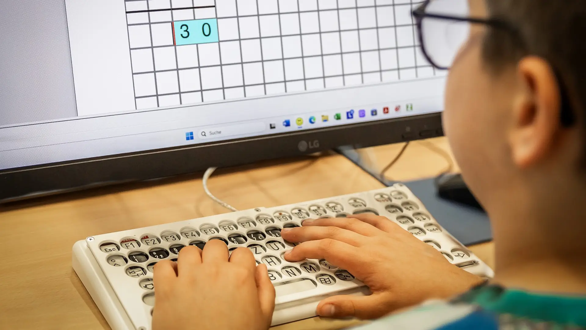 Ein Schüler sitzt am Schreibtisch und löst Rechenaufgaben. Er blickt dabei zum Monitor eines Computers und tippt an einer Blindentastatur.