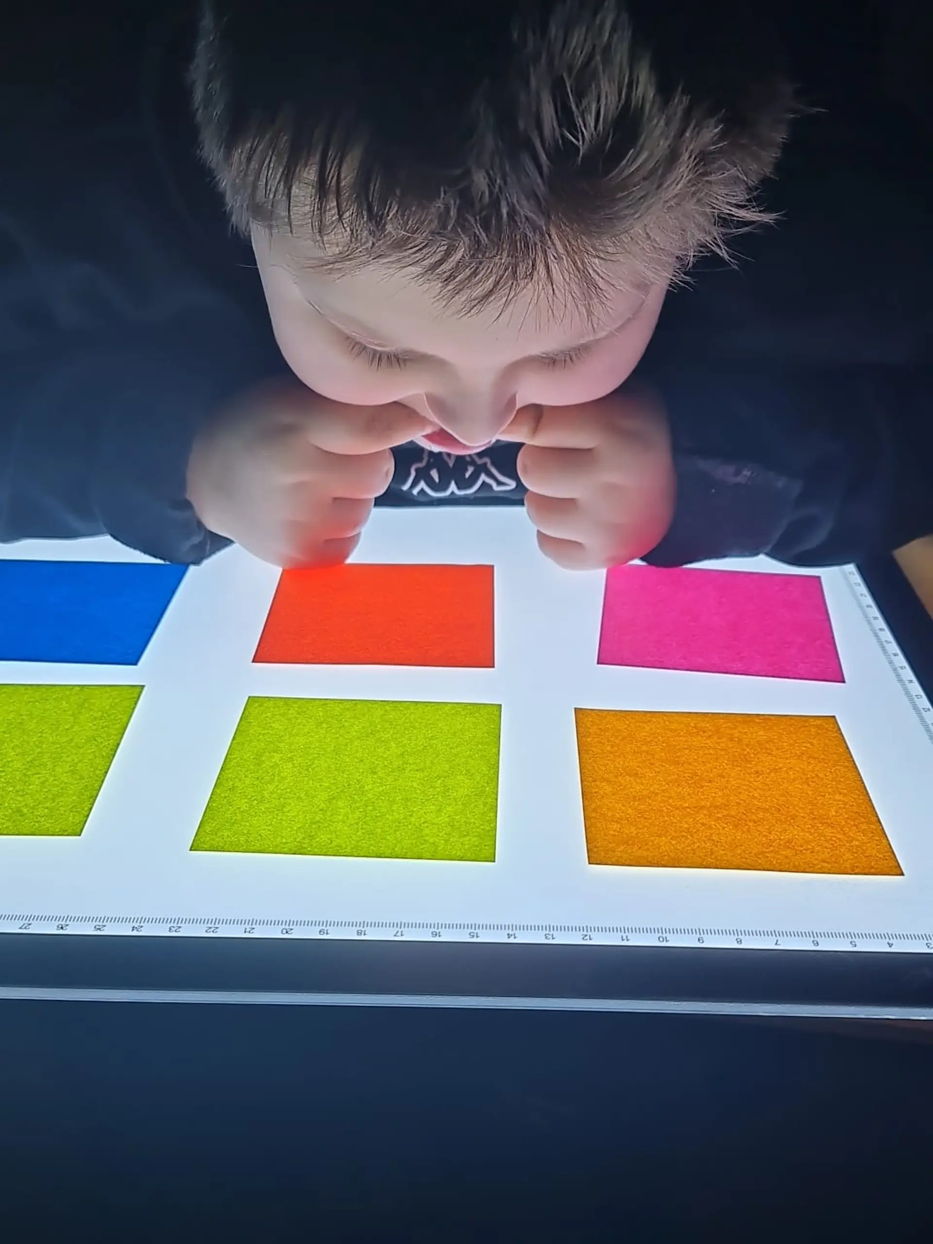 Ein Junge mit komplexer Beeinträchtigung sitzt ganz nah an einem Lichttisch. Auf der Oberfläche liegen sechs quadratische Papierblätter in verschiedenen Farben. Das Kind schaut sich diese konzentriert an und beißt dabei an seinen Zeigefingern.