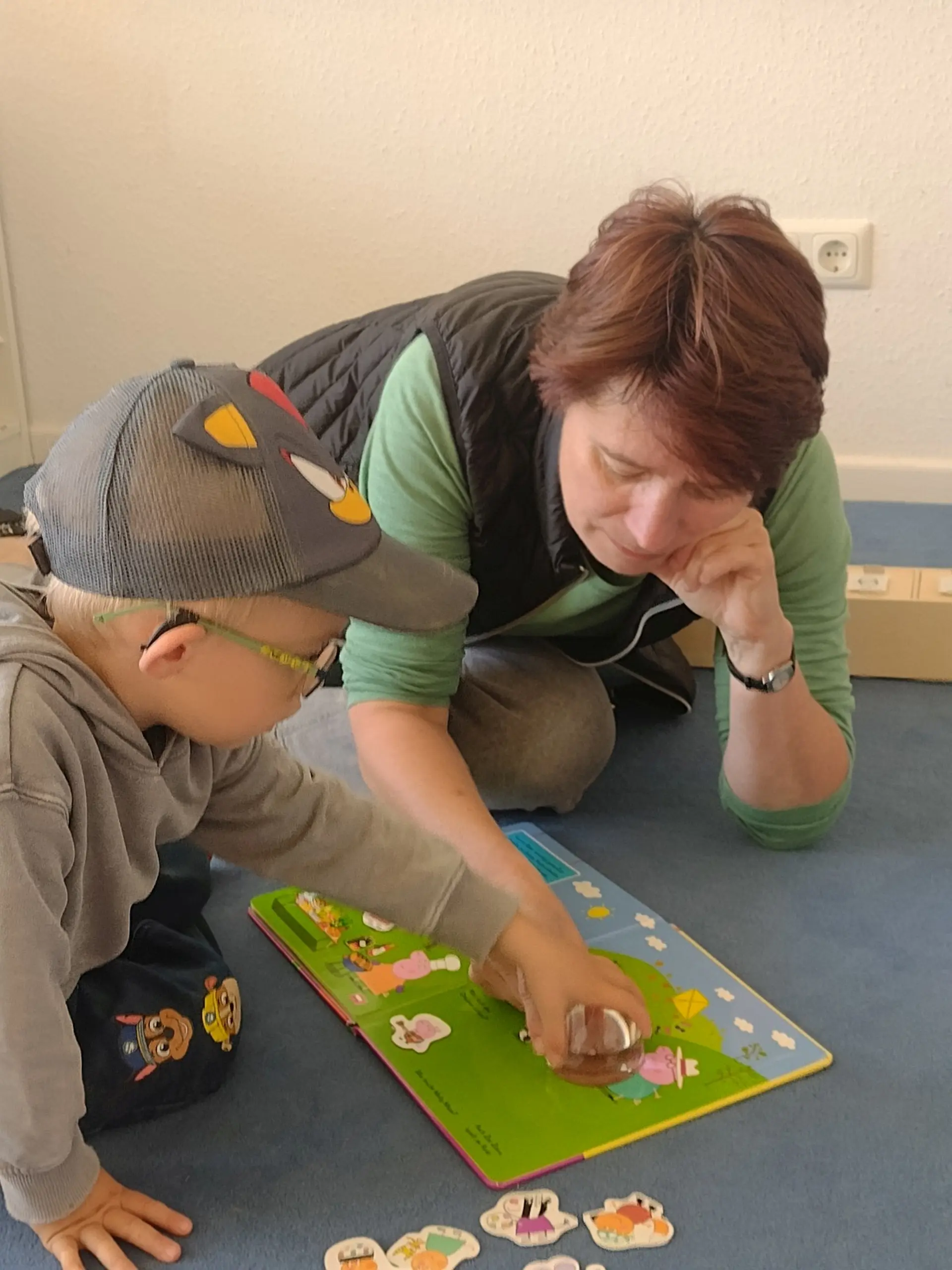 Eine Frühförderin und ein Kind mit Brille sitzen auf einem Teppichboden und schauen sich ein Peppa-Wutz-Bilderbuch an. Um die Bilder besser zu sehen, benutzt das Kind eine Vergrößerungslupe. Die Frühförderin hilft ihm dabei, diese zur richtigen Stelle zu verschieben.