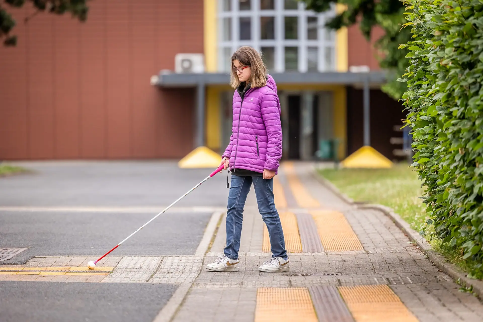 Eine Schülerin mit Blindenstock kommt aus dem Hauptgebäude heraus. Mit ihrem Stock ertastet sie den Weg dank der Markierungen auf dem Gehweg. Im Hintergrund ist das Schulgebäude unscharf zu sehen.