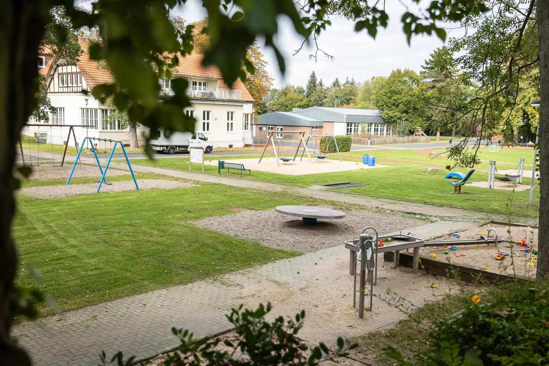 Ansicht eines Teils des Schulgeländes. Im Vordergrund ist ein großer Spielplatz mit verschiedenen Spielmöglichkeiten wie einem Sandkasten und einer Wasserspielanlage sowie vielen Schaukeln. Im Hintergrund ist ein Schulgebäude zu sehen.