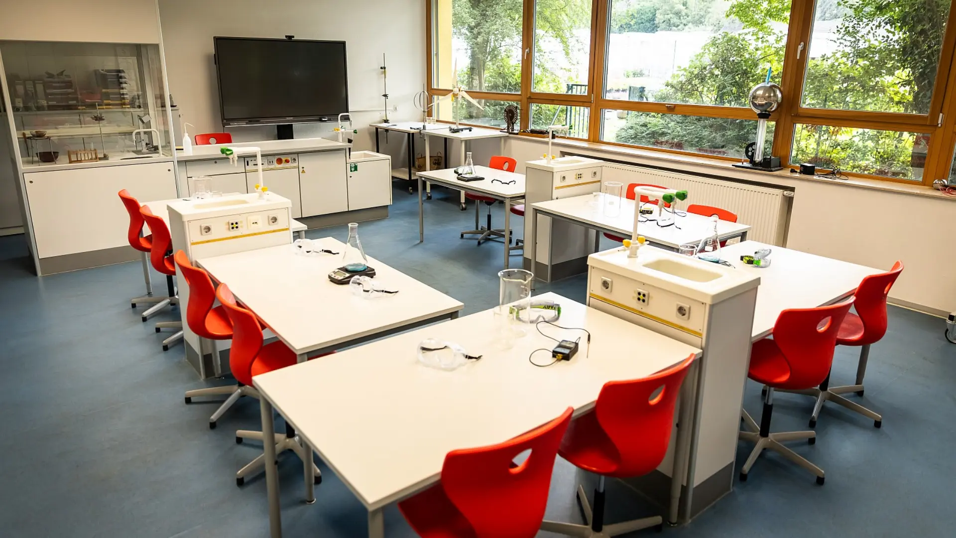 Mehrere Labortische mit integrierten Spülbecken und Wasserhähnen sind im Fachraum verteilt, umgeben von leuchtend roten Stühlen. Jeder Tisch ist mit verschiedenen Laborgeräten und Utensilien für Experimente ausgestattet. Im Hintergrund befindet sich ein großer Flachbildschirm an der Wand und eine Vitrine mit weiteren Geräten. Große Fensterfronten lassen viel Tageslicht herein und bieten einen Blick auf Bäume.