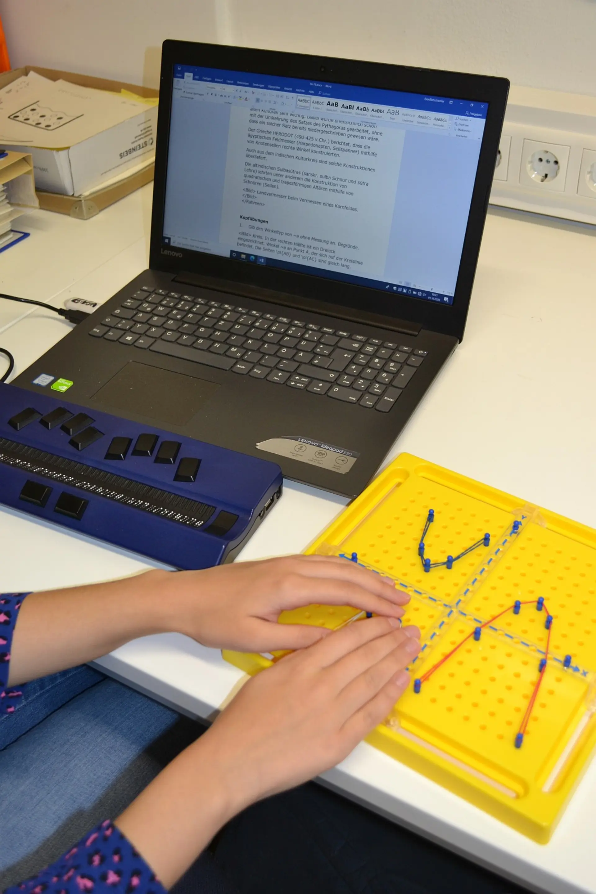 Die Hände einer Schülerin, die an ihrem Laptoparbeitsplatz mit Braillezeile sitzt und auf dem danebenliegenden Koordinatensteckbrett einen Graphen abtastet. 