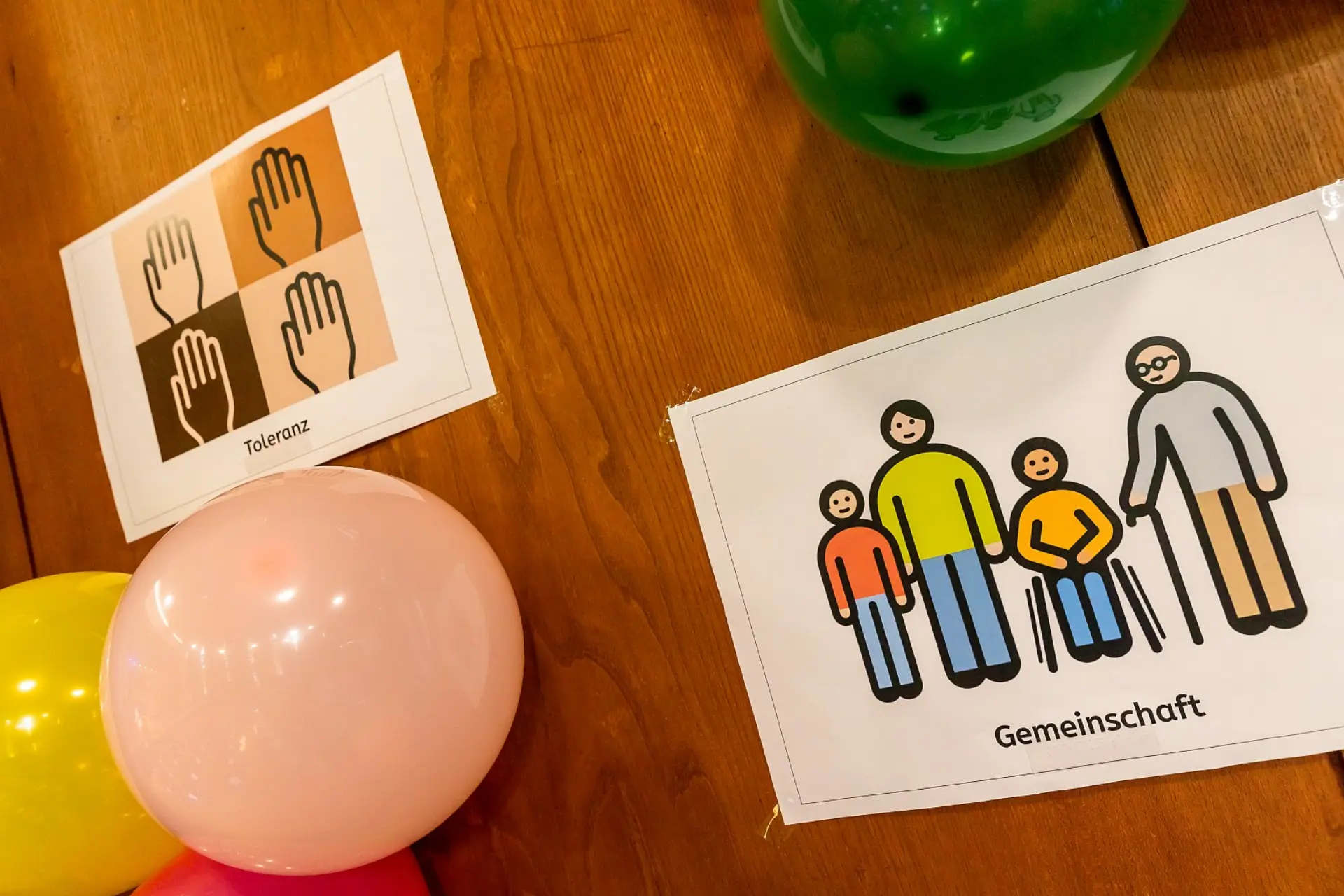 Eine Holzwand in der Aula, die mit Ballons und Blättern mit Metacom-Symbolen darauf beschmückt ist. Auf den beiden Blättern in diesem Bild sind die Symbole für Toleranz und Gemeinschaft zu sehen.