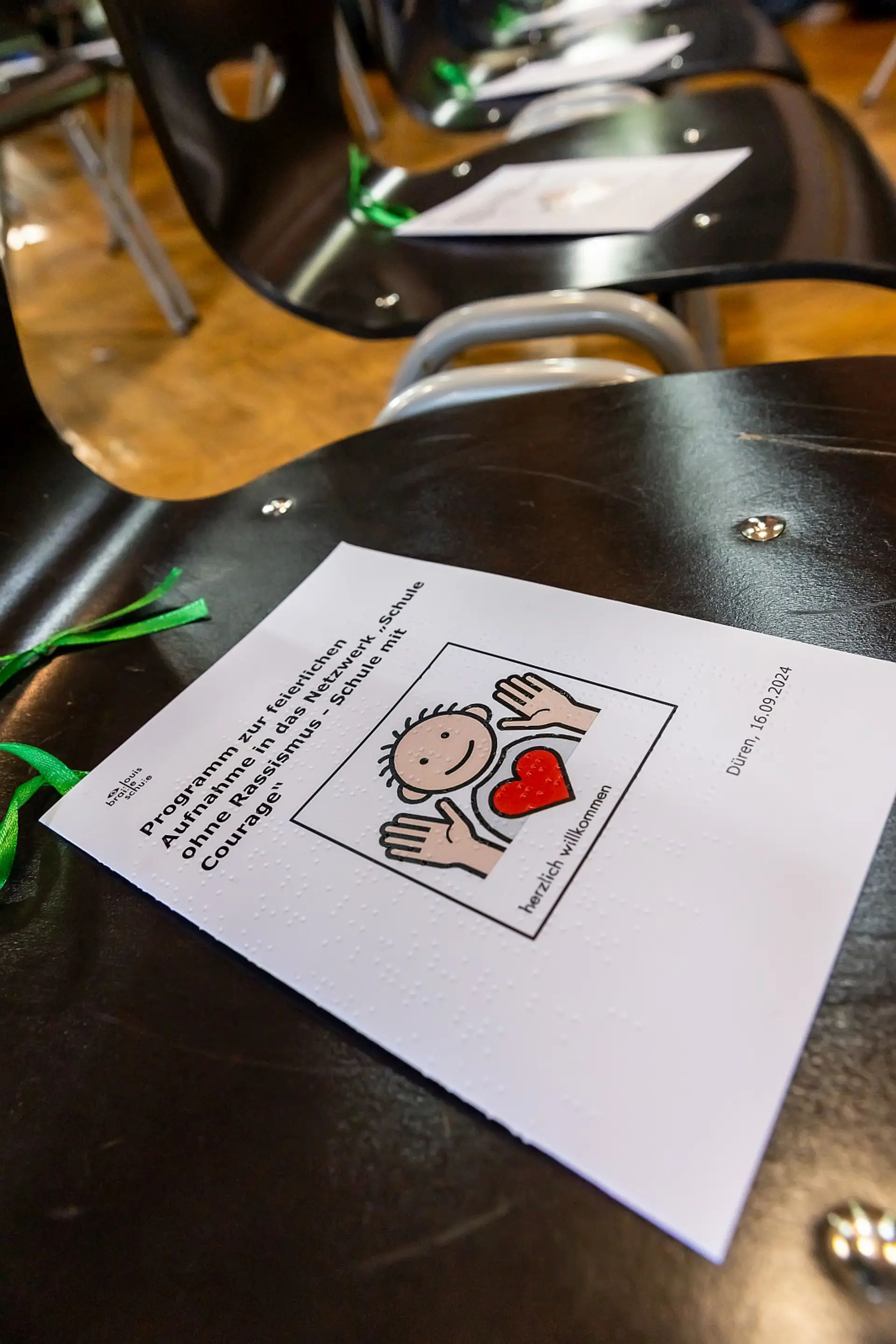 Eine Reihe mit Stühlen in der Aula. Auf dem Stuhl im Vordergrund liegt das Festprogramm. In Textform und in Braille-Schrift steht auf dem Cover: Programm zur feierlichen Aufnahme in das Netzwerk 'Schule ohne Rassismus, Schule mit Courage. Düren, 16.9.2024'. Dazu sieht man auf dem Cover noch das 'Herzlich willkommen'-Metacom-Symbol.