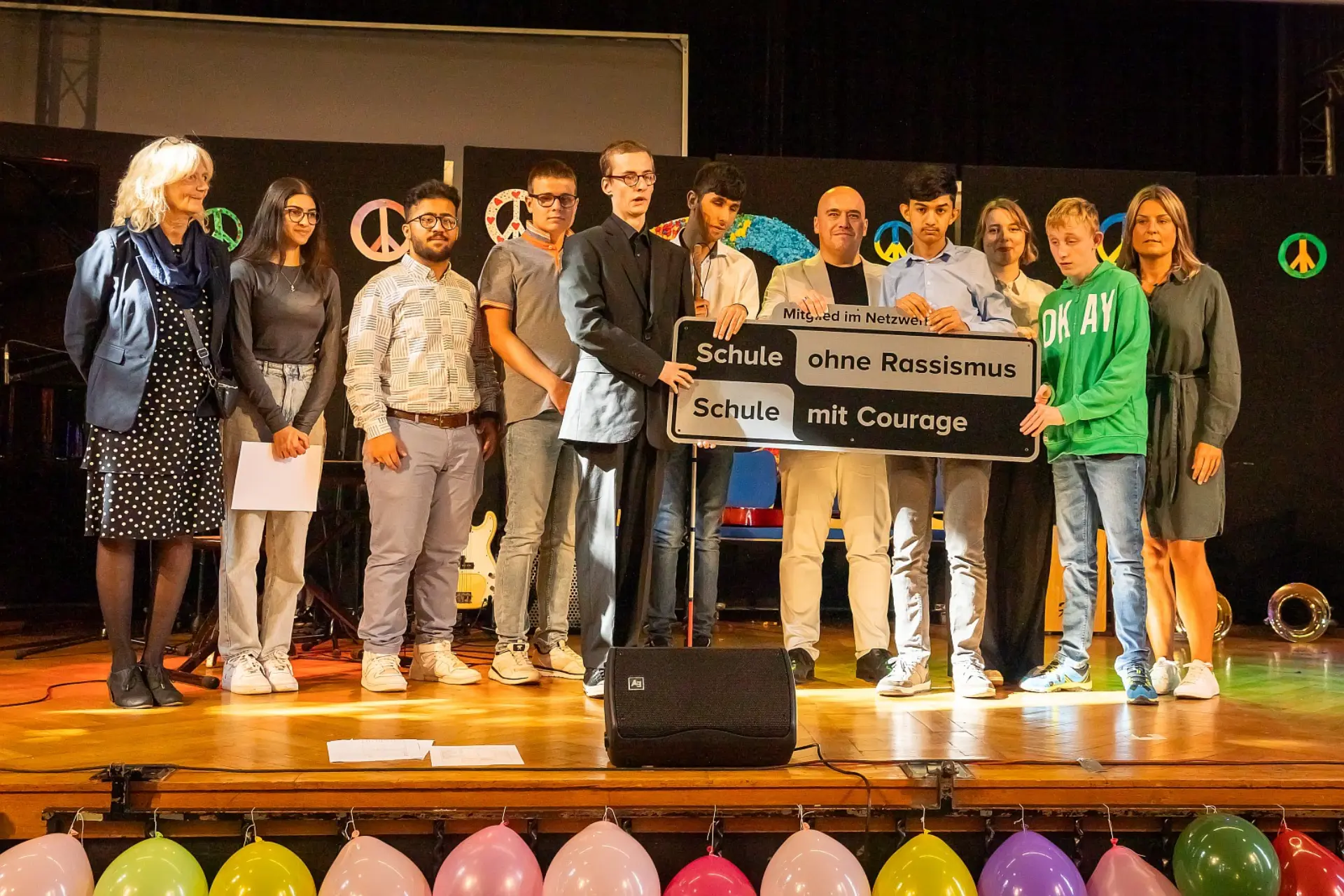 Ein Gruppenbild von Schüler*innen der Abschlussstufe sowie Erwachsenen stehen auf der feierlich geschmückten Bühne der Schulaula. Die Personen in der Mitte halten das Schild 'Mitglied im Netzwerk: Schule ohne Rassismus, Schule mit Courage'.