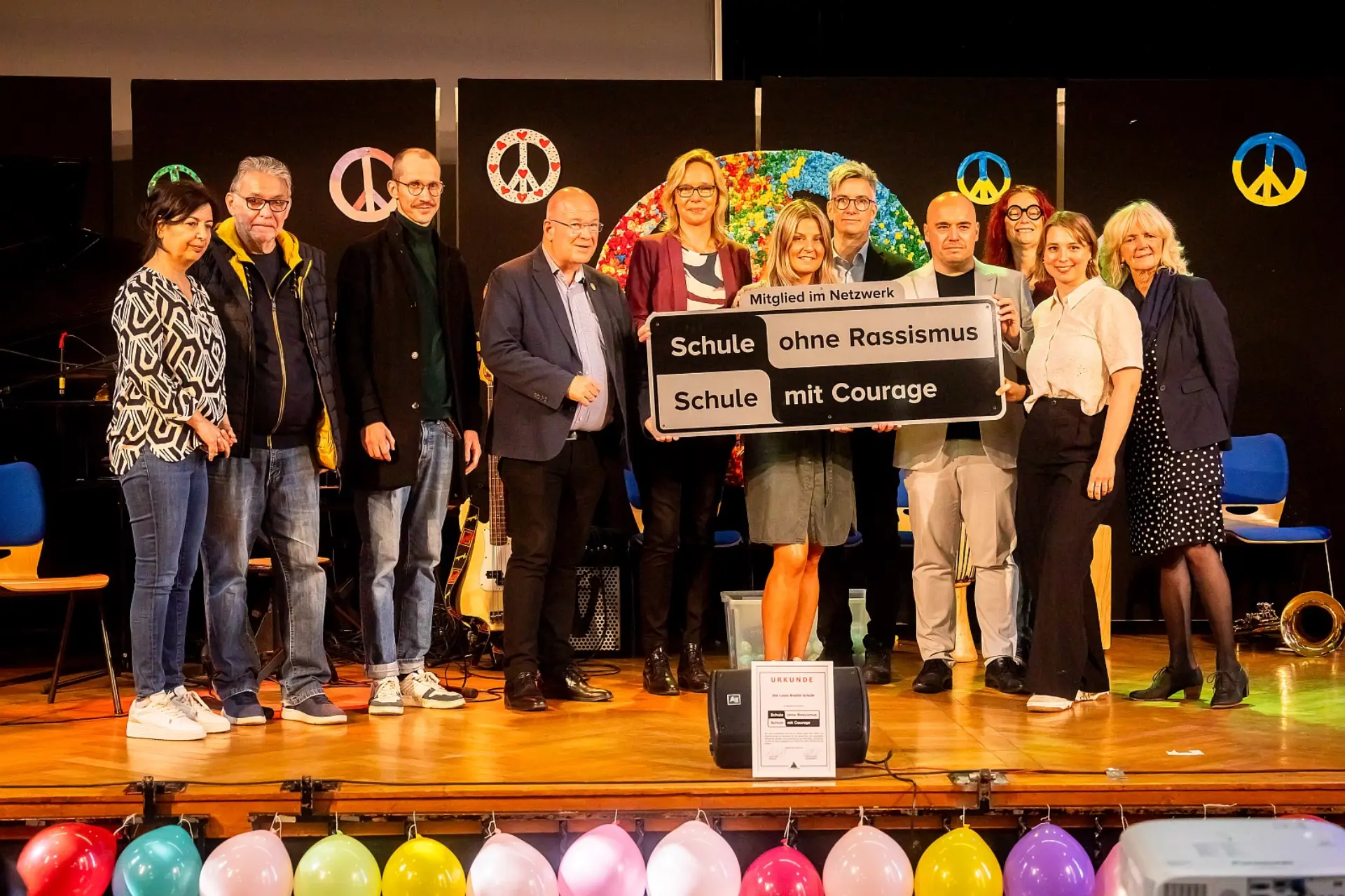 Ein Gruppenbild von elf Erwachsenen, die auf der Bühne der Aula stehen. Die Person in der Mitte hält das Schild ' Mitglied im Netzwerk: Schule ohne Rassismus, Schule mit Courage'. Vor ihnen auf dem Boden ist die gleichnamige Urkunde für die Schule an einem Lautsprecher angelehnt.