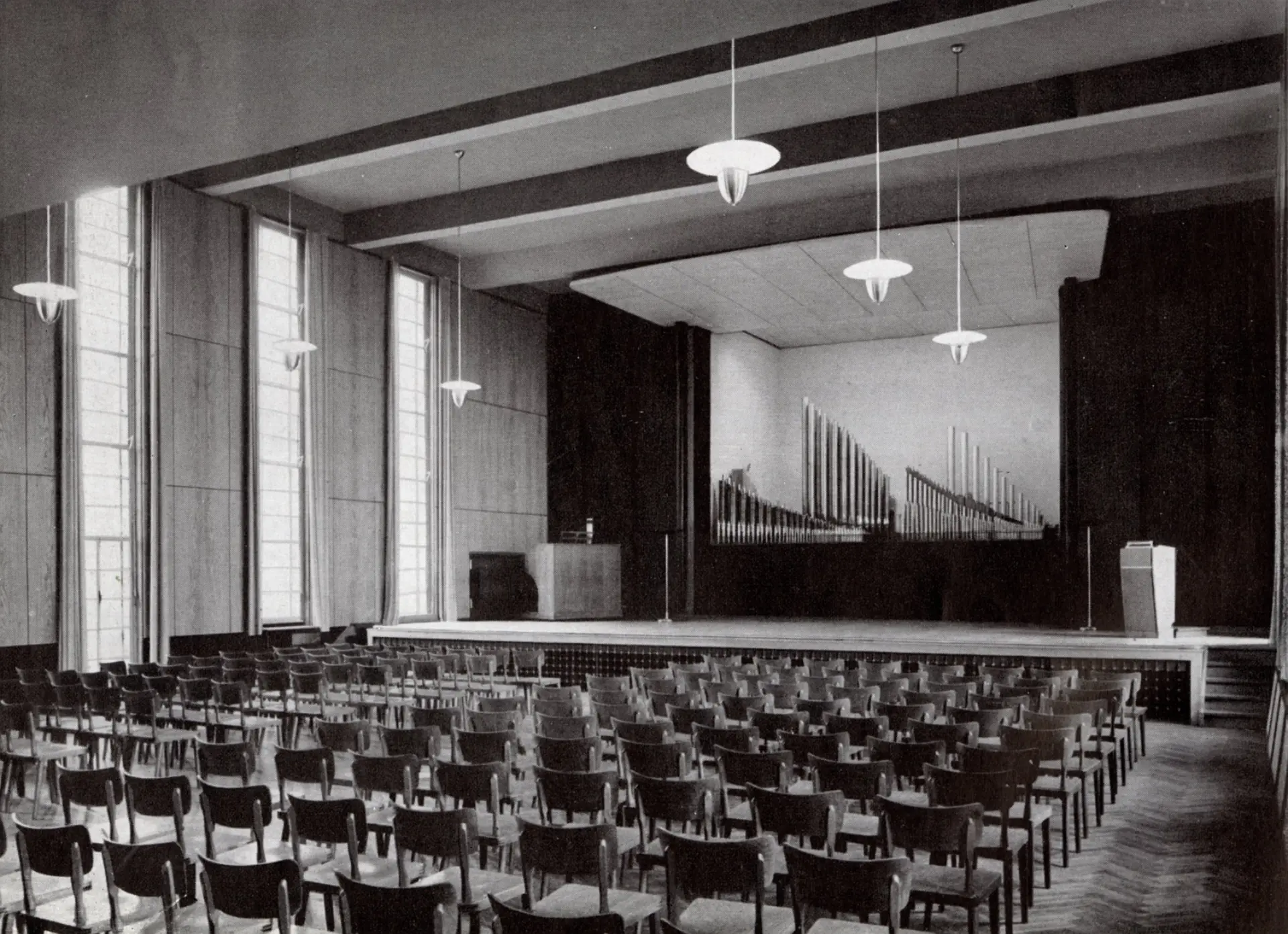 Ein monochromes Foto zeigt das Innere einer leeren Aula. Der Raum ist mit Reihen von Stühlen ausgestattet, die auf einen erhöhten Bühnenbereich ausgerichtet sind. Auf der Bühne befindet sich im Hintergrund eine große Orgel mit sichtbaren Pfeifen. Die Wände des Saales sind mit Holz vertäfelt und auf der linken Seite befinden sich hohe, schmale Fenster, die natürliches Licht hereinlassen. Von der Decke hängen mehrere moderne Pendelleuchten herab. Das gesamte Ambiente wirkt schlicht und funktional, was auf eine Bauweise aus der Mitte des 20. Jahrhunderts hindeutet.