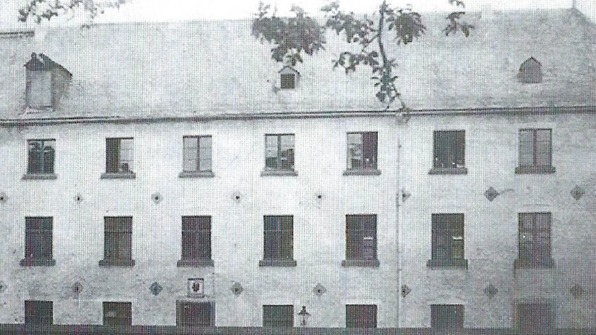 Ein monochromes, historisches Foto zeigt das ehemalige Jesuitenkloster, das um 1910 als Blindenanstalt genutzt wurde. Das Gebäude ist ein mehrstöckiges, rechteckiges Bauwerk mit zahlreichen Fenstern, die in Reihen angeordnet sind. Das Dach ist steil und besitzt mehrere Dachgauben. Die Fassade weist eine schlichte Gestaltung auf, wobei im unteren Bereich des Gebäudes ein Wappen oder Emblem zu erkennen ist.