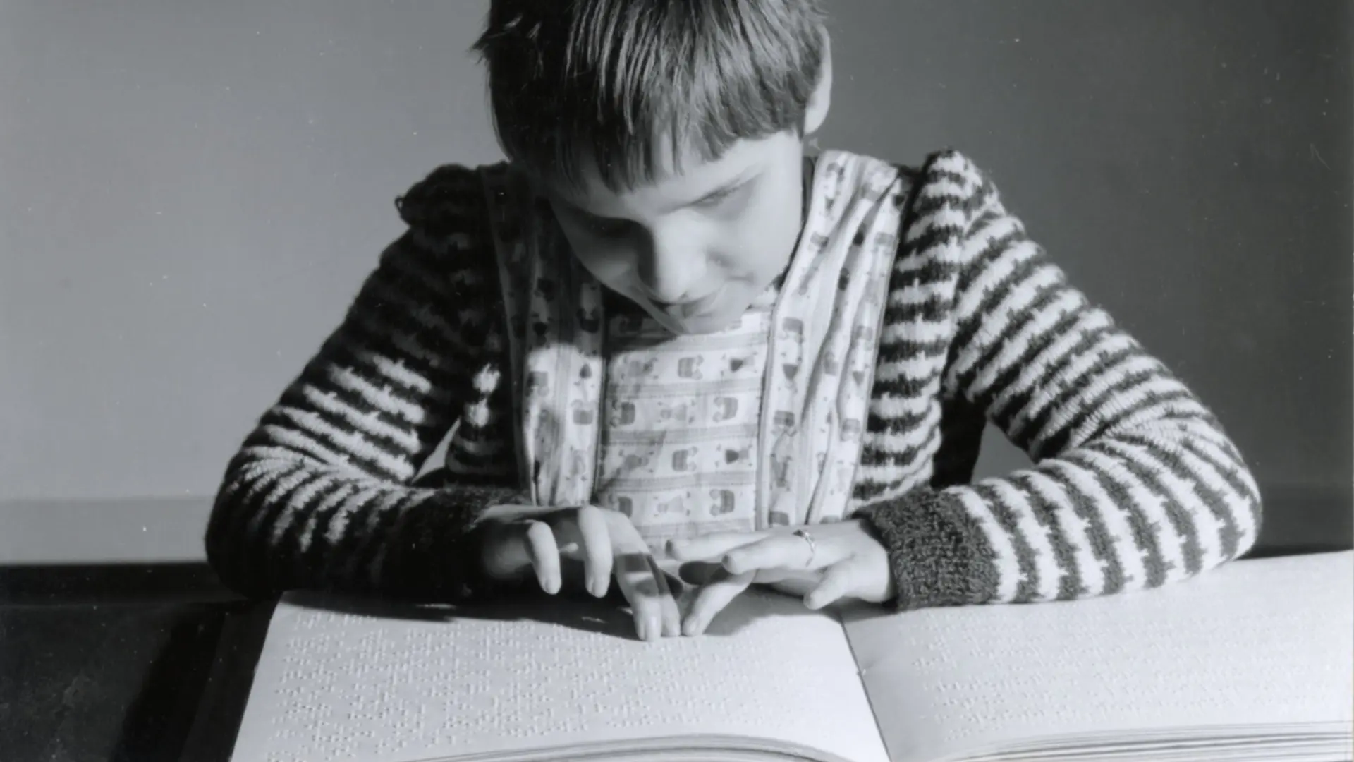 Ein monochromes Foto von einer blinden Schülerin, die am Tisch sitzt. Vor ihr ist ein Buch in Braille-Schrift, sie geht mit den Fingern darüber und liest den Text.