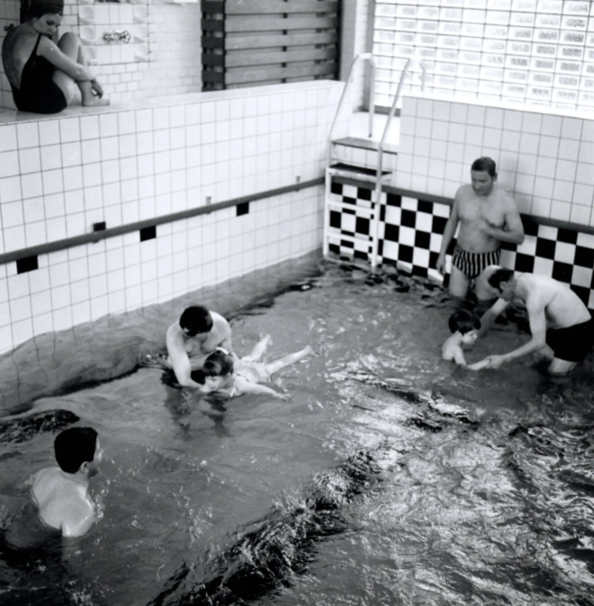 Eine schwarzweiße Aufnahme vom Schwimmunterricht in der Blindenanstalt. Zu sehen ist ein Wasserbecken mit mit niedrigem Wasser, die Wände drum-herum sind mit Fliesen bedeckt. Im Wasser sind vier Männer in Badehosen, zwei davon helfen jeweils einem Schulkind beim Schwimmen oder sich im Wasser bewegen. Eine Frau im Badeanzug steht am Rand des Schwimmbeckens und beobachtet das Geschehen.