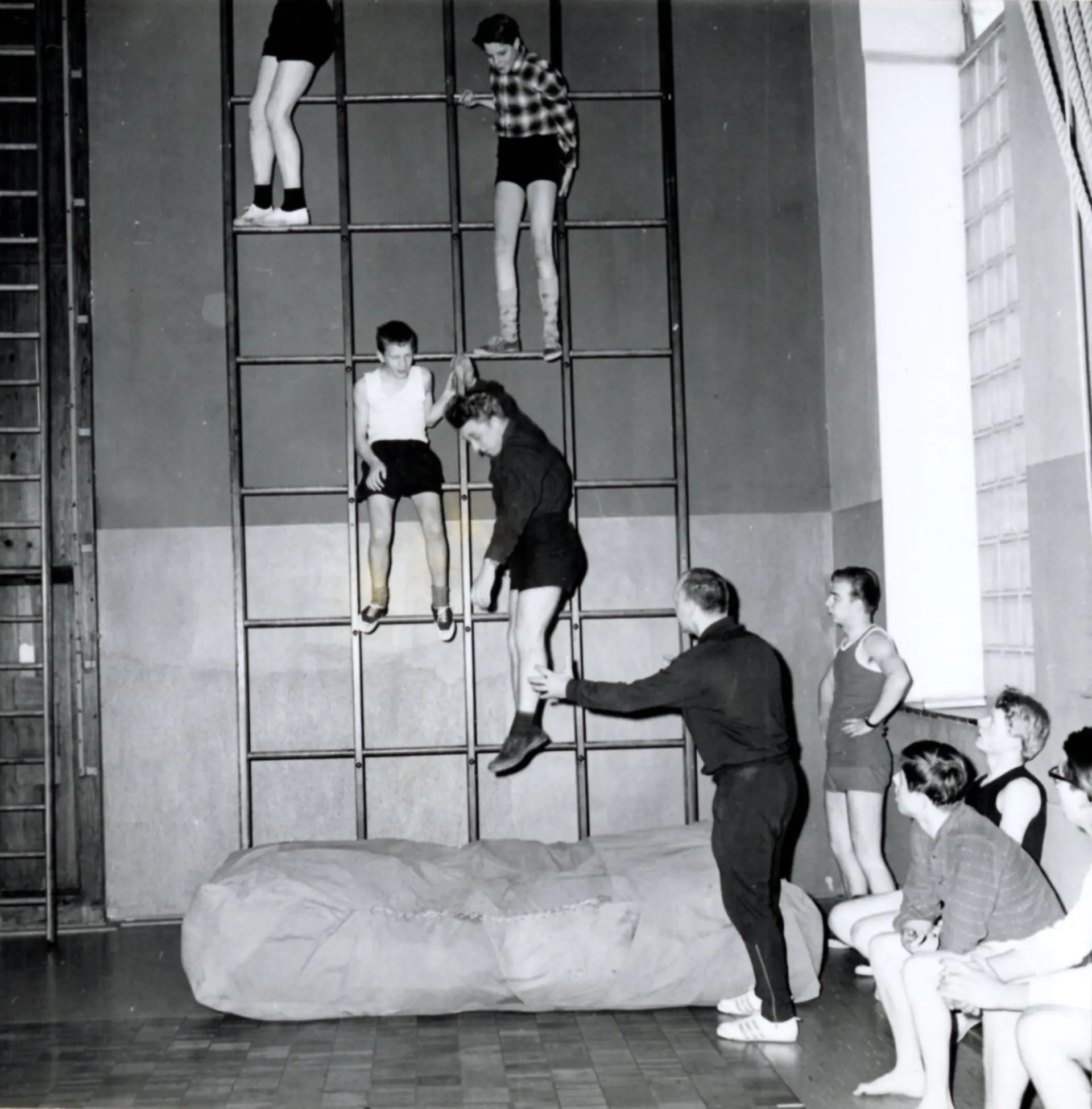 Eine Schwarzweiß-Aufnahme aus der Sporthalle der Schule. Hier ist die Kletterwand mit Holzsprossen, direkt darunter eine dicke Schutzmatratze zu sehen. Vier Schüler sind hochgeklettert und befinden sich auf verschiedenen Höhen, einer davon springt gerade auf die Matratze herunter. Eine Lehrer im Sportanzug steht vor der Matratze und streck seinen Arm zum Jugendlichen, um ihn aufzuhalten, falls es seitlich hinfällt. Vier andere Jugendliche in Sportsbekleidung stehen oder sitzen an der Seite und beobachten das Geschehen.