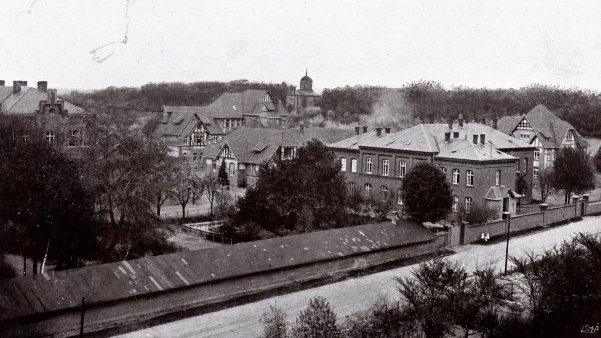 Ein historisches Schwarz-Weiß-Luftbild zeigt eine Gesamtansicht eines weitläufigen Gebäudekomplexes in einer ländlichen oder vorstädtischen Umgebung. Im Vordergrund verläuft eine Straße parallel zum unteren Bildrand, daneben befindet sich ein langes, niedriges Gebäude mit einem Schrägdach. Dahinter erstrecken sich mehrere größere Gebäude mit verschiedenen Dachformen, darunter Sattel- und Walmdächer, die von Bäumen und anderen Grünflächen umgeben sind. Im Hintergrund ist ein dichter Wald oder Baumbestand zu sehen, aus dem sich eine Kirchenkuppel oder ein Turm erhebt.