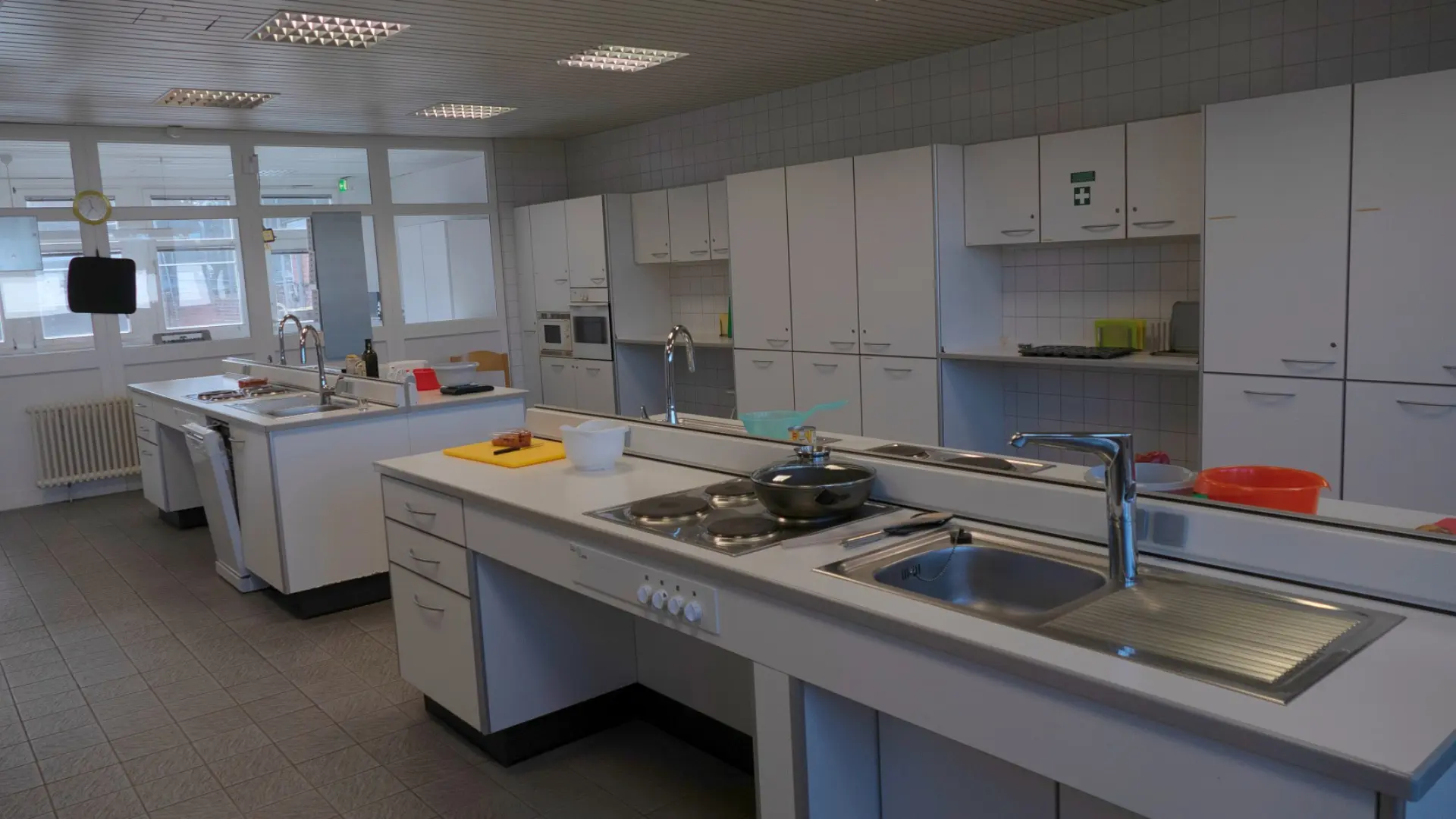 Ein gut beleuchteter, sauberer Raum, der als Lehrküche oder Hauswirtschaftszentrum dient. Es gibt mehrere weiße Küchenzeilen mit Spülbecken und Kochfeldern. An den Wänden sind viele Hängeschränke angebracht. Eine große Fensterfront auf der linken Seite lässt Tageslicht herein. Auf den Arbeitsflächen stehen verschiedene Küchenutensilien und Kochgeschirr. Der Boden ist mit Fliesen ausgelegt.