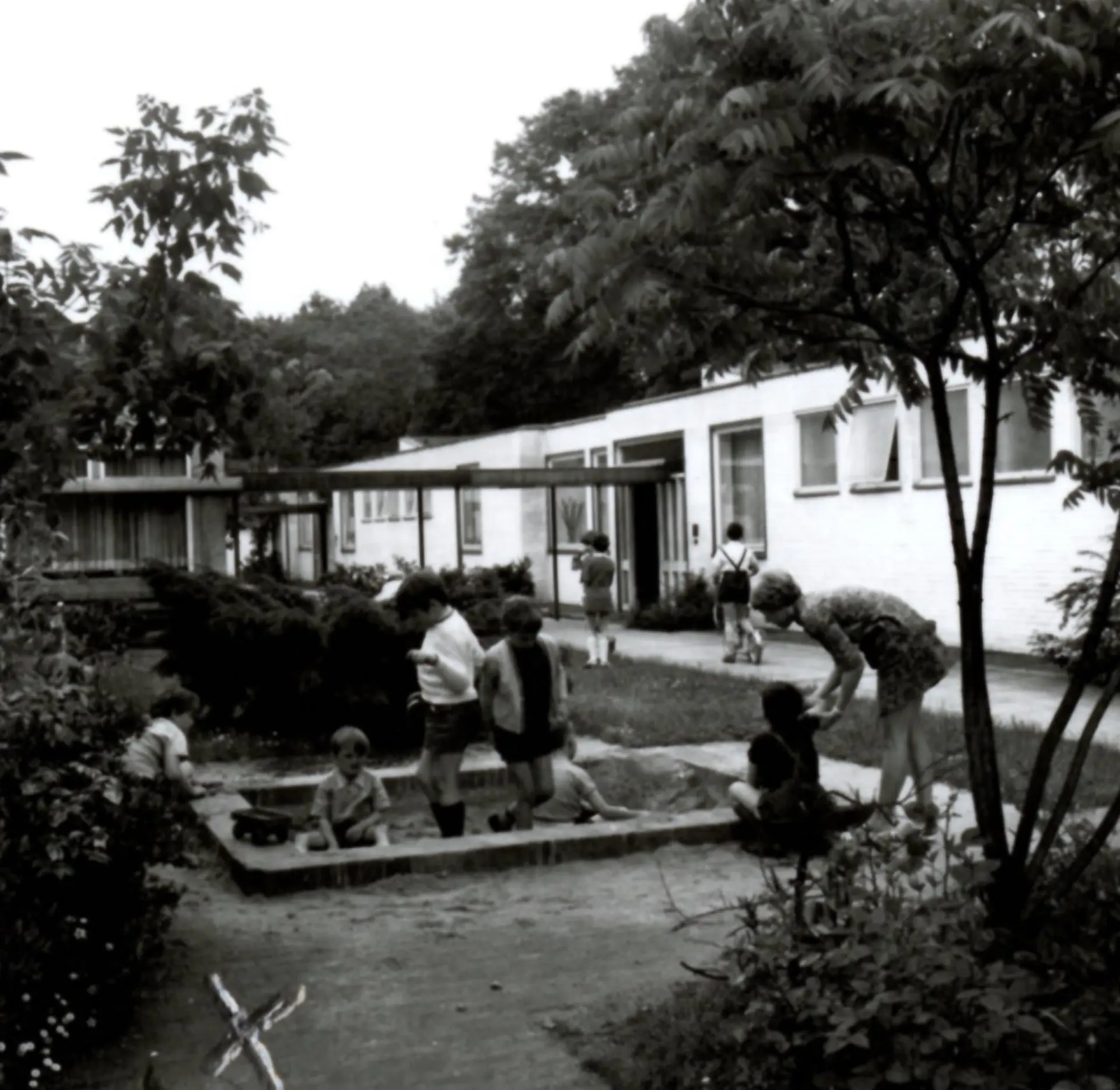 Eine Schwarzweiß-Aufnahme, die einen kleinen Spielplatz mit einem quadratischen Sandkasten auf dem Schulgelände zeigt. Sech Schüler*innen spielen im und um den Sandkasten, eine Lehrkraft beugt sich zu einem der Kinder. Im Hintergrund ist ein flaches, einstöckiges Gebäude mit vielen Fenstern zu sehen. Davor stehen noch zwei Schüler*innen, auf dem asphaltierten Gehweg davor ist ein Kind mit Tretroller. Drum-herum sind viele Büsche und Bäume mit vielen Blättern zu sehen. Dies und die Kleidung der Schüler*innen zeigt, dass es Sommer ist. 
