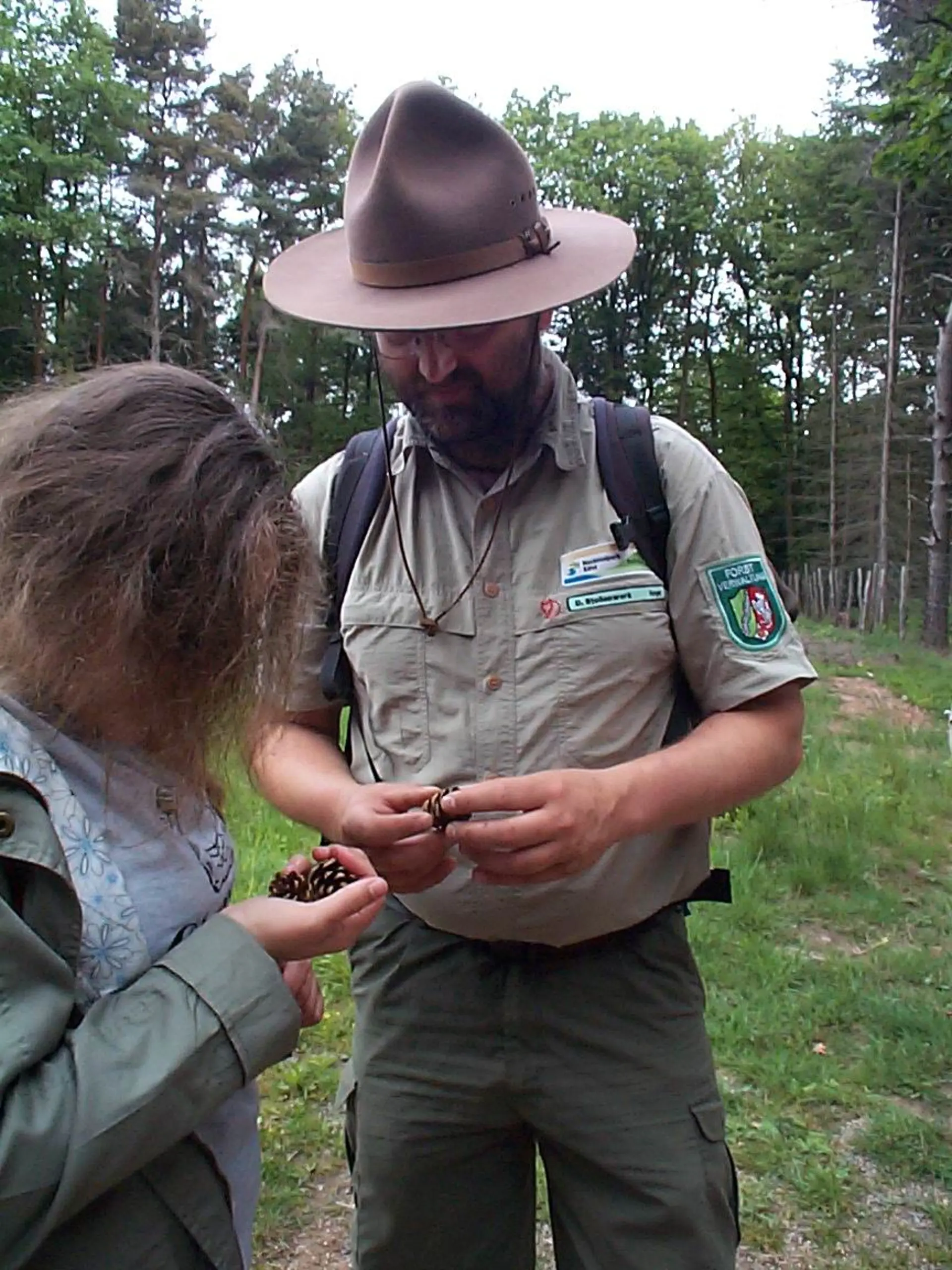 Ein Mann mit Bart, einem Ranger-Hut und Ranger-Klamotten hält in seinen Händen einen Tannenzapfen und schaut auf ihn herab. An seinem Hemd sind mehrere Abzeichen befestigt, darunter eines mit dem Schriftzug "Nationalpark Eifel" und eines mit dem Wort "Forstverwaltung" und dem Wappen von Nordrhein-Westphalen. Eine Schülerin steht links vor ihm und hält ebenfalls einen Tannenzapfen in ihren Händen. Sie befinden sich in einem Waldgebiet im Nationalpark Eifel, das ist auch an den großen Tannenbäumen im Hintergrund und dem teilweise bewachsenen Boden erkennbar.