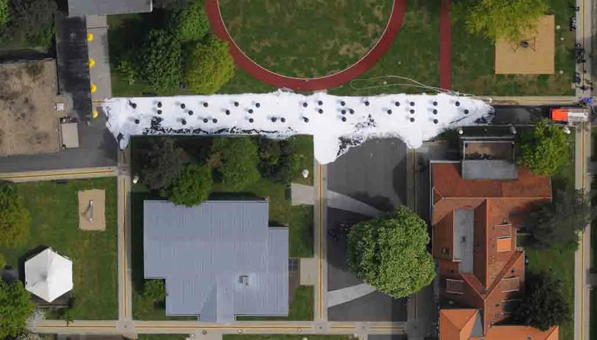 Luftaufnahme mit einer Drohne: Darauf ist das Schulgelände mit drei Schulgebäuden, drei Sandkästen sowie Grasflächen und frischen Bäumen zu sehen. Der breite, gradlinige Gehweg in der Mitte der Bildkomposition ist komplett weiß, da er mit Löschschaum gedeckt worden ist. Darauf sind schwarze Punkte aus den Regenschirmen der Schulkinder zu sehen, die den Schriftzug "Louis Braille" in Braille-Schrift darstellen. 