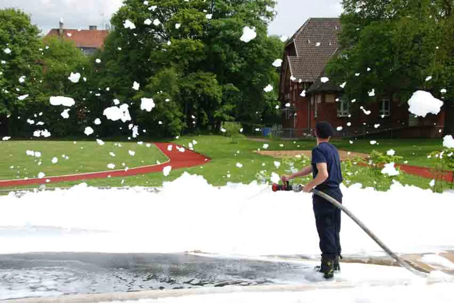 Ein Mann in Feuerwehr-Kleidung sprüht aus einem Schlauch große Mengen weißen Schaum auf den Boden des Außengeländes der Schule. Überall in der Luft und auf dem umliegenden Gras schweben Schaumflocken. Im Hintergrund sind grüne Bäume, ein Laufweg und ein Gebäude zu sehen.