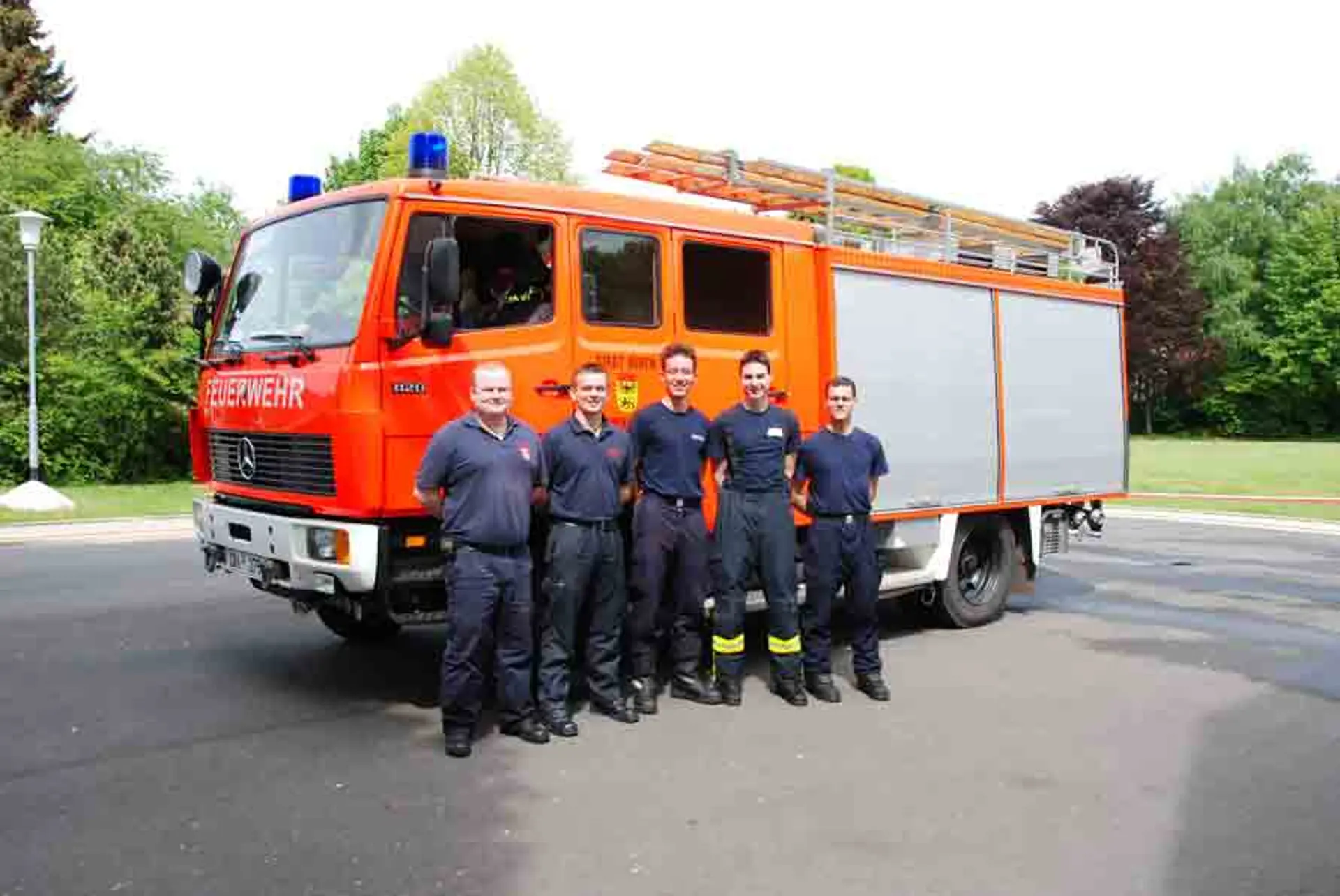 Ein Feuerwagen ist auf einer asphaltierten Fläche auf dem Schulgelände geparkt. Davon stehen fünf Feuerwehrmänner, die in die Kamera schauen und lächeln. Im Hintergrund sind Bäume mit frischem Laub zu sehen.