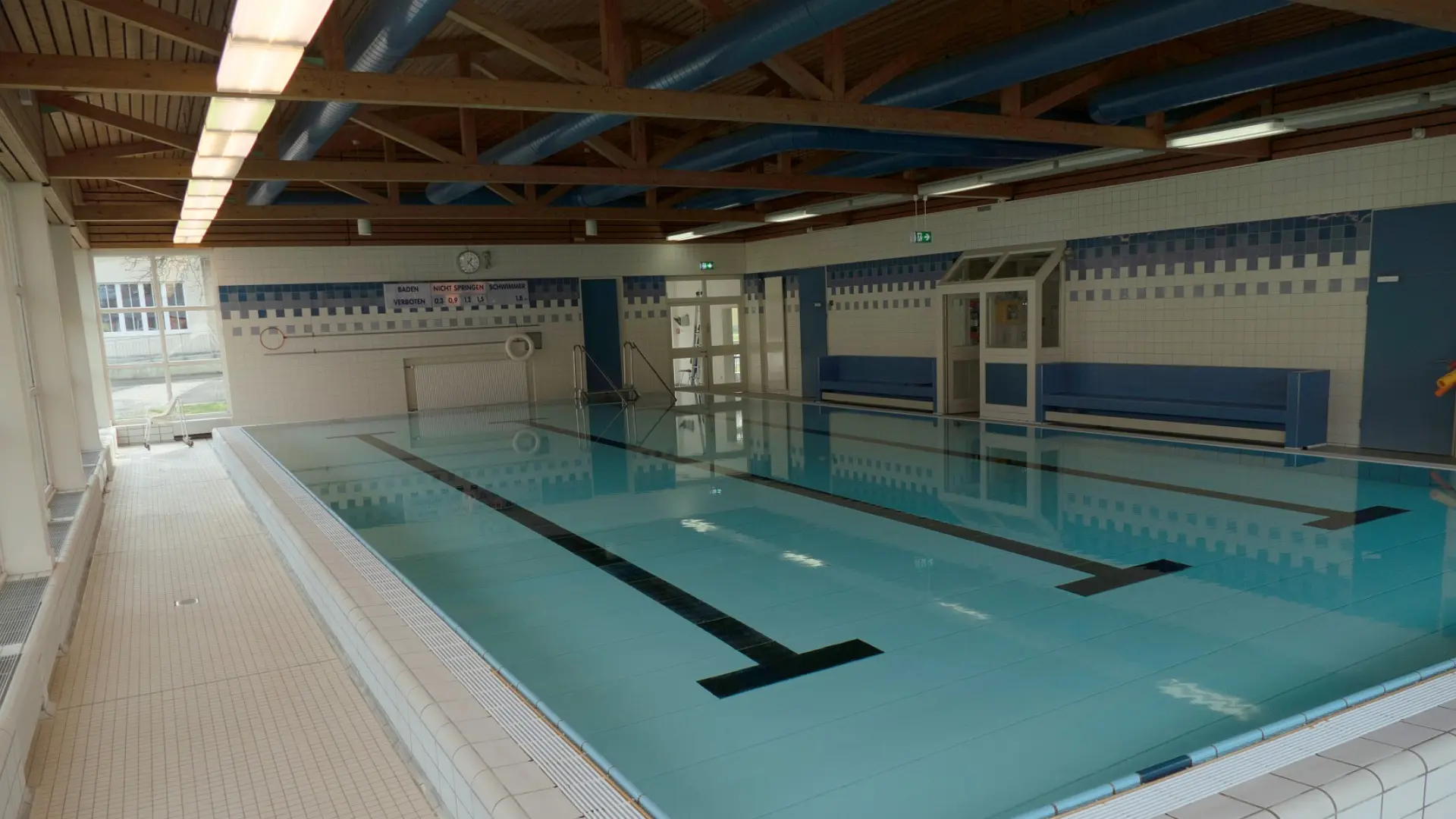 Ein Schwimmbad-Innenraum, von einer erhöhten Position aus fotografiert. Das Becken ist mit klarem Wasser gefüllt und hat zwei dicke Linien, die vom Startblock bis zum anderen Ende verlaufen. Die Wände sind gefliest. Im Hintergrund sind Fenster, Bänke und Türen zu sehen. Die Decke besteht aus Holzbalken.