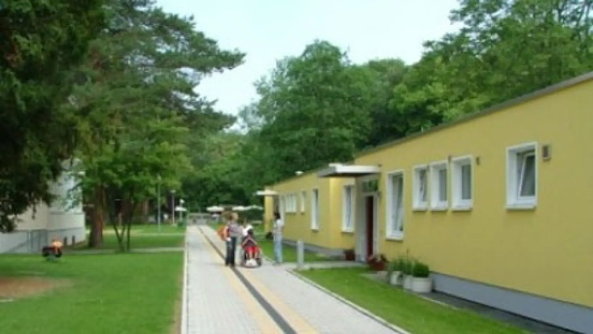 Das Internatsgebäude der LVR-Louis-Braille-Schule von draußen. Dieses befindet sich rechts im Bild. Es ist ein flacher Bau, nur mit Erdgeschoss und mit mehreren Eingängen und vielen Fenster der Zimmer. Davor ist ein breiter, betonierter Fußweg mit taktilen Markierungen zur Orientierung. Vor dem einen Eingang stehen drei Personen, die sich unterhalten, eine davon schiebt noch eine Person im Rollstuhl. Links im Bild befindet sich ein anderes Schulgebäude. Links und rechts hinter dem Internat sind viele Bäume mit grünen Baumkronen zu sehen.