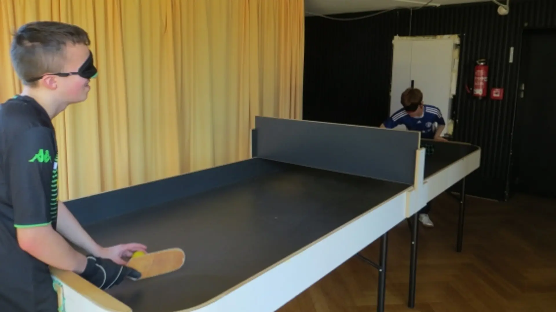 Zwei Schüler spielen Tischball in einem Raum. Der Raum hat keine Fenster auf der Seite, wo der Tisch steht, sondern ein schwerfallender Vorhang. Hinter dem einen Jungen ist auch der Ausgang.