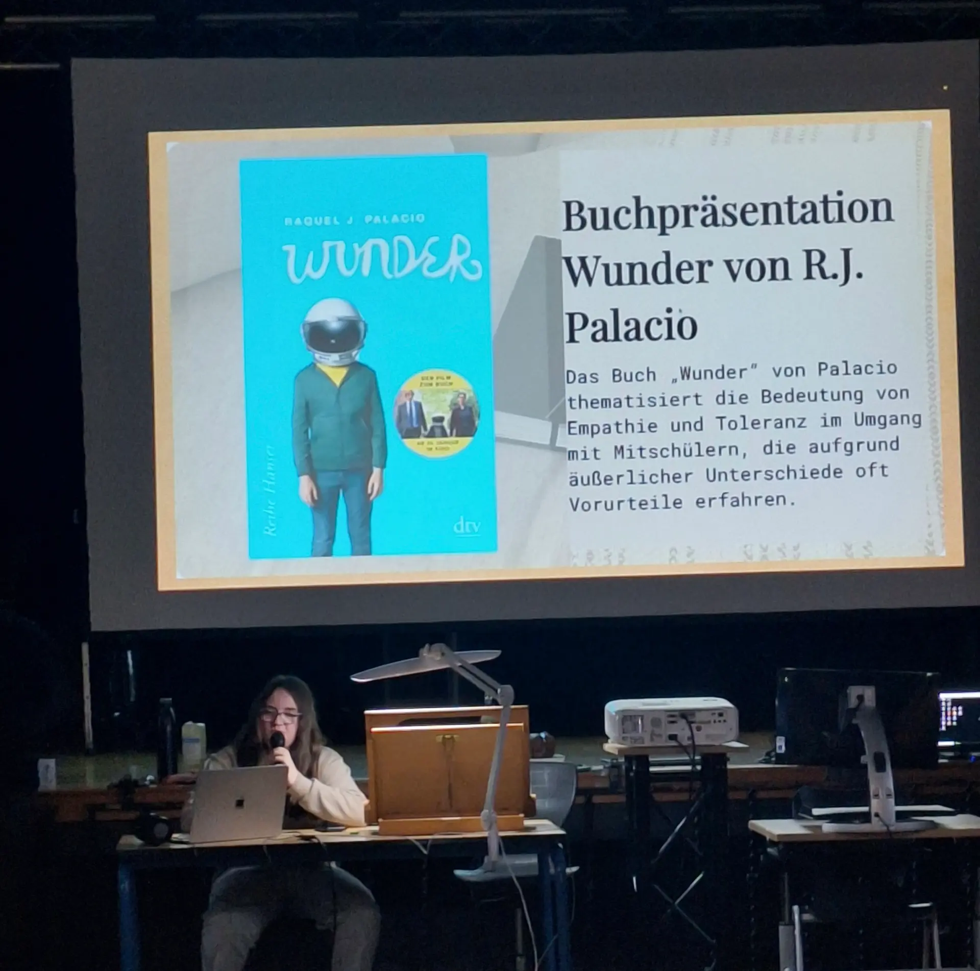 Eine Schülerin sitzt vorne in der Aula an einem Tisch. Ein Laptop vor ihr und ein Mikro in der Hand. Hinter ihr ist eine Leinwand zu sehen, auf die die Folie einer PowerPoint-Präsentation projiziert ist, die das Cover des Buches „Wunder“ sowie eine kurze Buchbeschreibung zeigt.