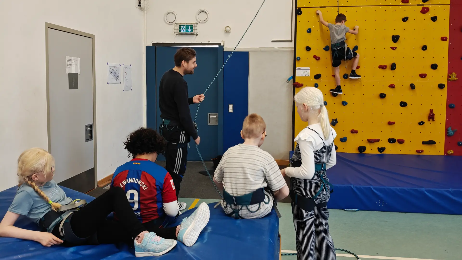 Ein Schüler klettert mit Sicherung an einer Kletterwand. Ein Lehrer hält das Sicherungsseil. Vier weitere Schüler*innen warten mit Klettergurt auf ihren Einsatz auf einer Weichbodenmatte.