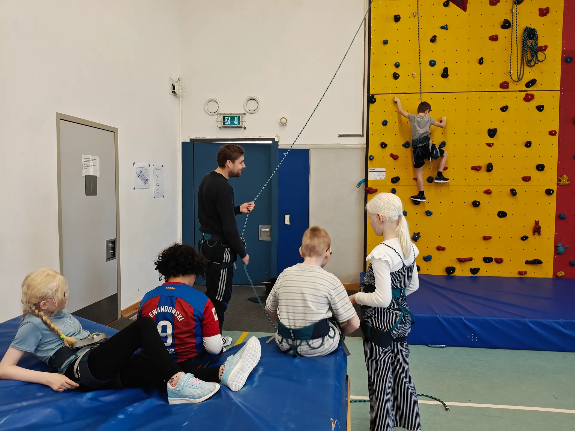 Ein Schüler klettert mit Sicherung an einer Kletterwand. Ein Lehrer hält das Sicherungsseil. Vier weitere Schüler*innen warten mit Klettergurt auf ihren Einsatz auf einer Weichbodenmatte.