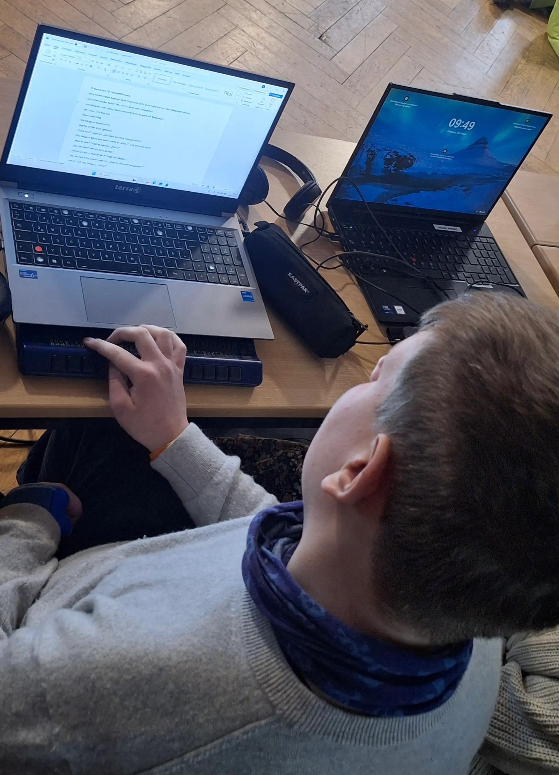 Ein Schüler sitzt an einem Tisch, vor sich einen Laptop mit Braillezeile. Er liest mit der rechten Hand auf der Braillezeile.