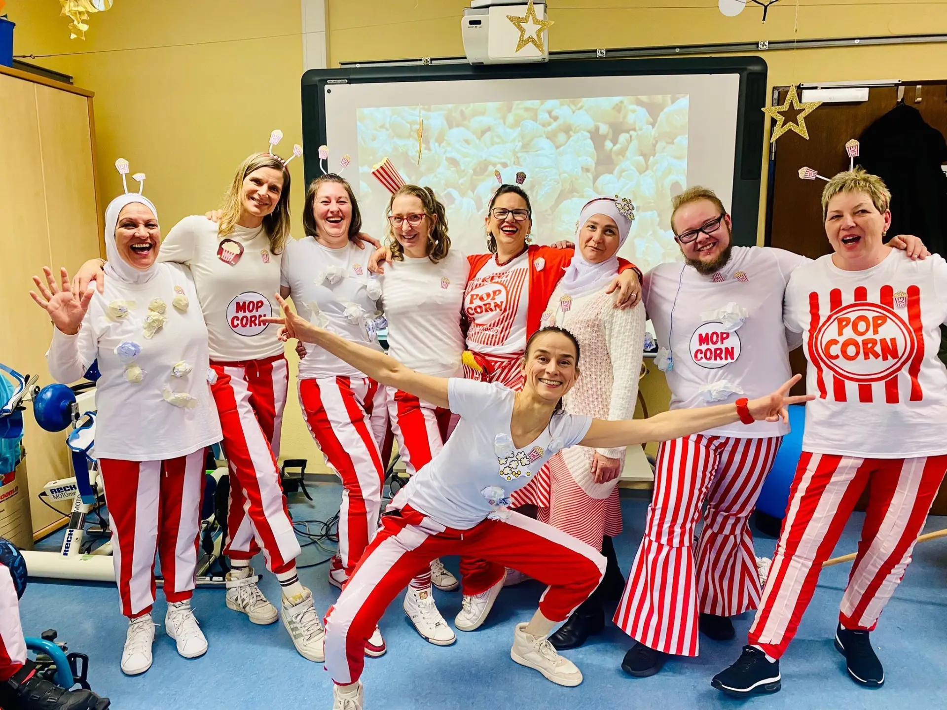 Auf dem Foto sind neun lachende und erwachsene Personen zu sehen. Alle sind als Popcorn verkleidet. Hinter den Personen ist eine digitale Tafel mit einem Popcorn-Bild zu sehen