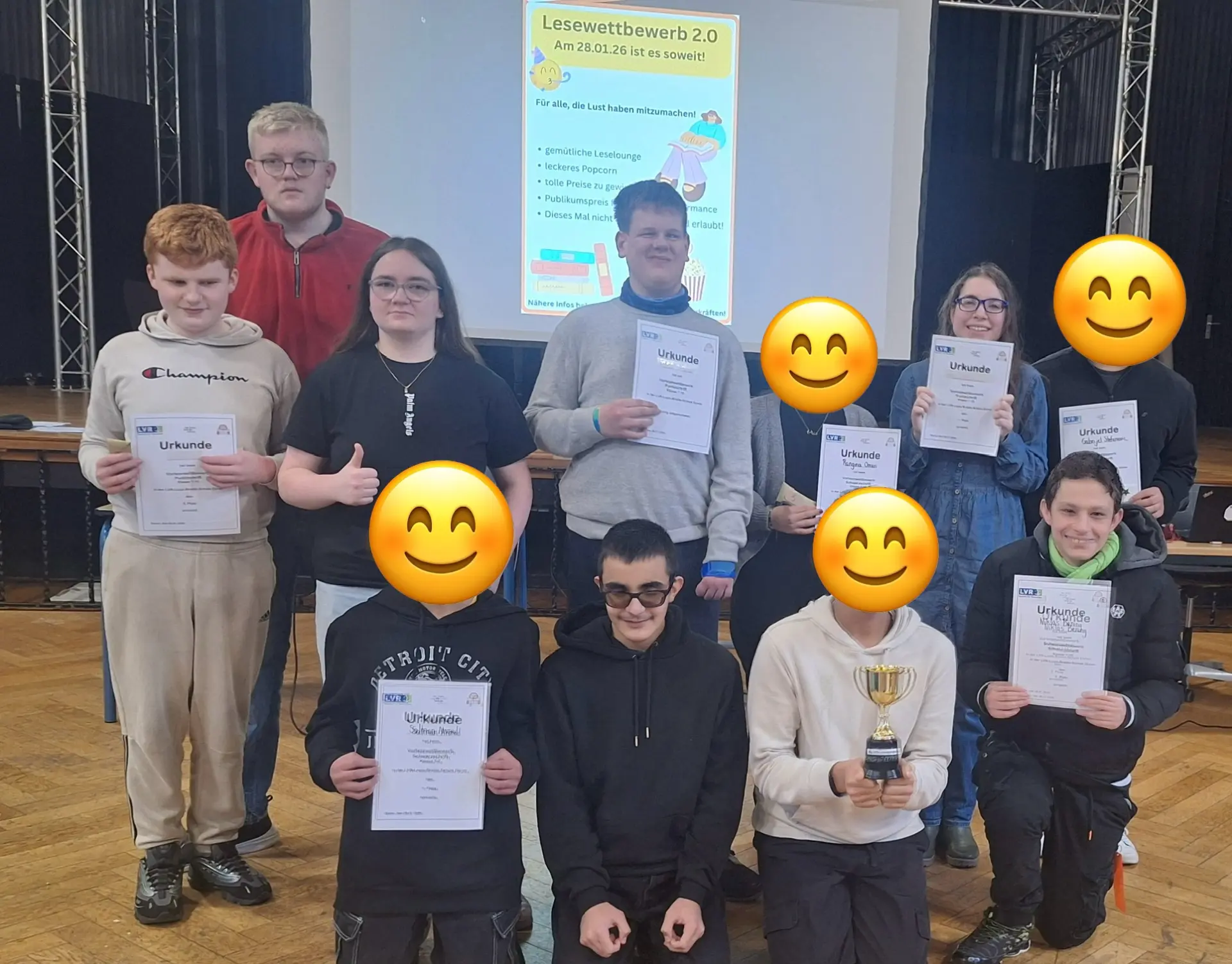 Auf dem Foto sind insgesamt sieben Schüler*innen, zu einem Gruppenfoto aufgestellt, zu sehen. Sie halten Urkunden sowie einen Pokal. Vier Gesichter sind mit Smileys abgedeckt. Im Hintergrund ist die Leinwand mit dem Plakat des Lesewettbewerbs zu sehen.