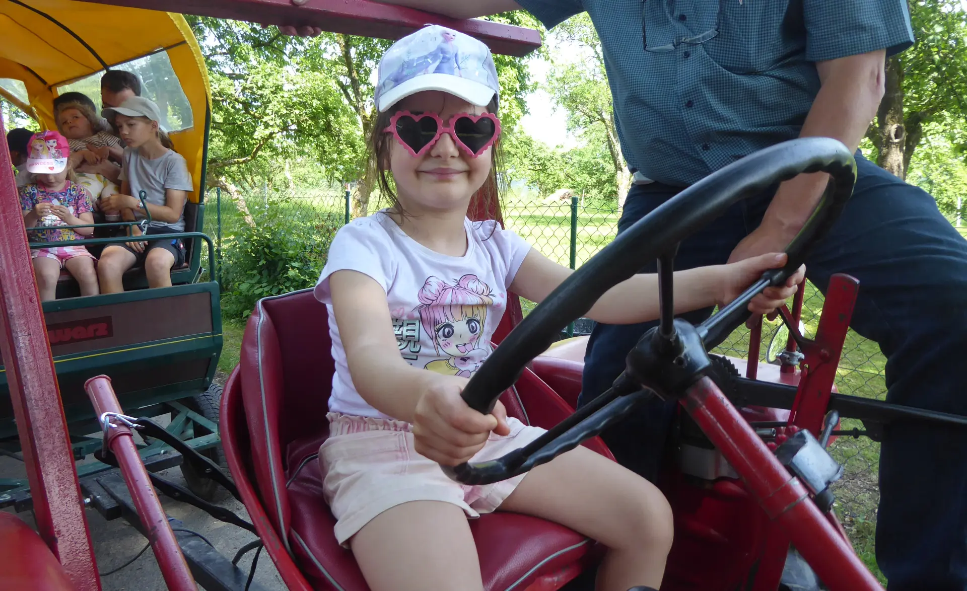 Ein Mädchen sitzt auf einem Traktor. Im Hintergrund sieht man die Familien im Planwagen.