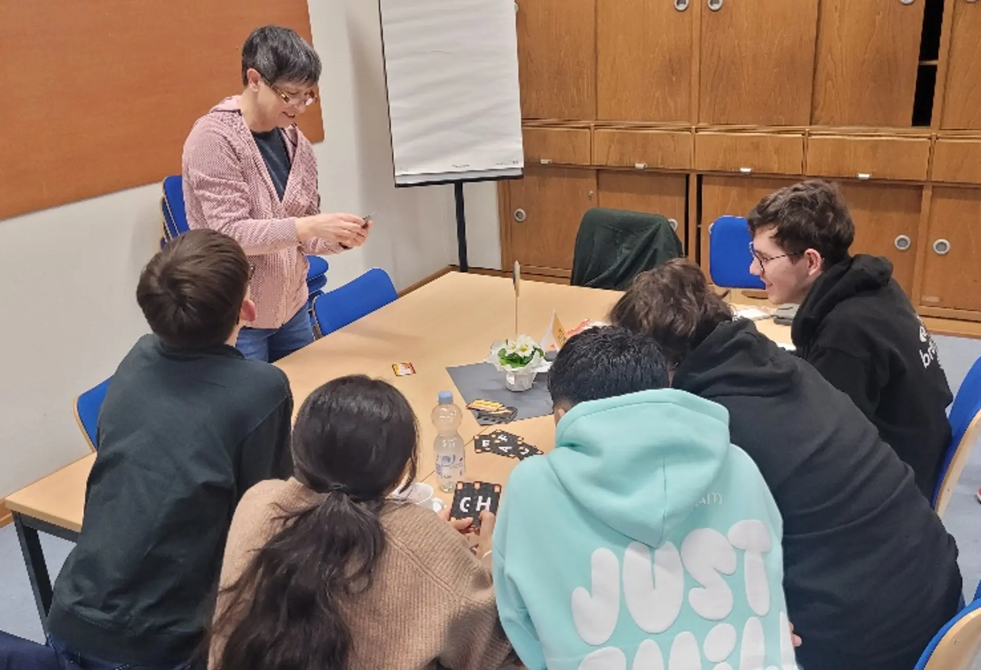 Eine Lehrerin steht an einem Tisch und erklärt interessierten Schüler*innen das Spiel.