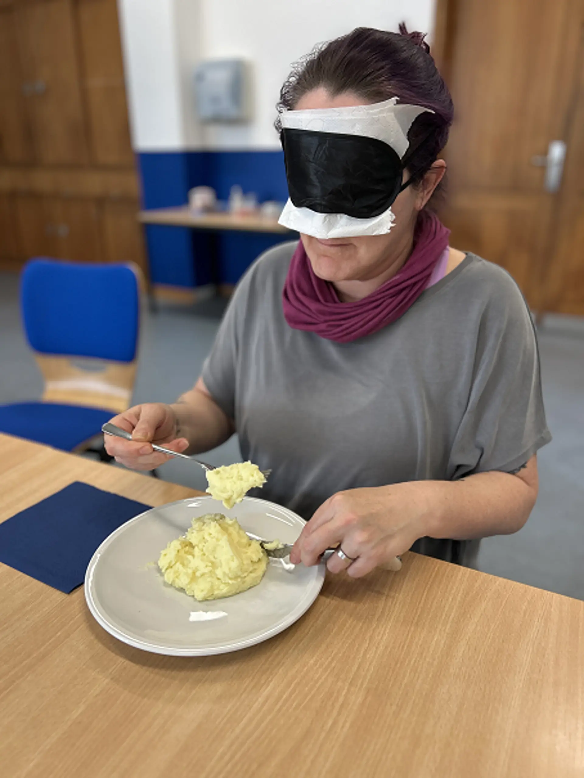 Eine Schulbegleiterin isst Kartoffelpüree mit Messer und Gabel unter der Augenbinde.