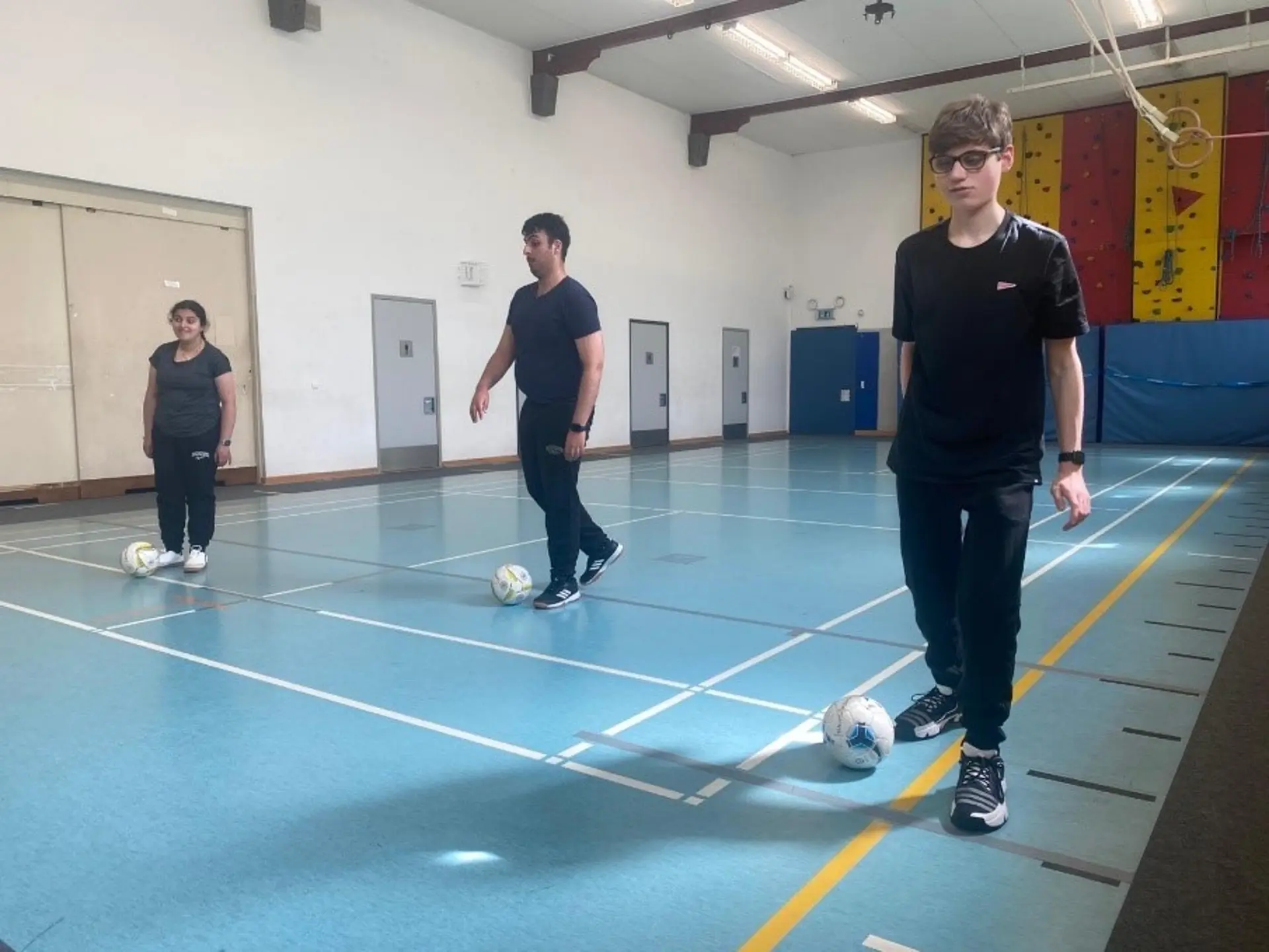 Drei Schüler*innen stehen in der Turnhalle. Vor ihnen liegt jeweils ein Ball und sie versuchen diesen gegen die Wand zu schießen.