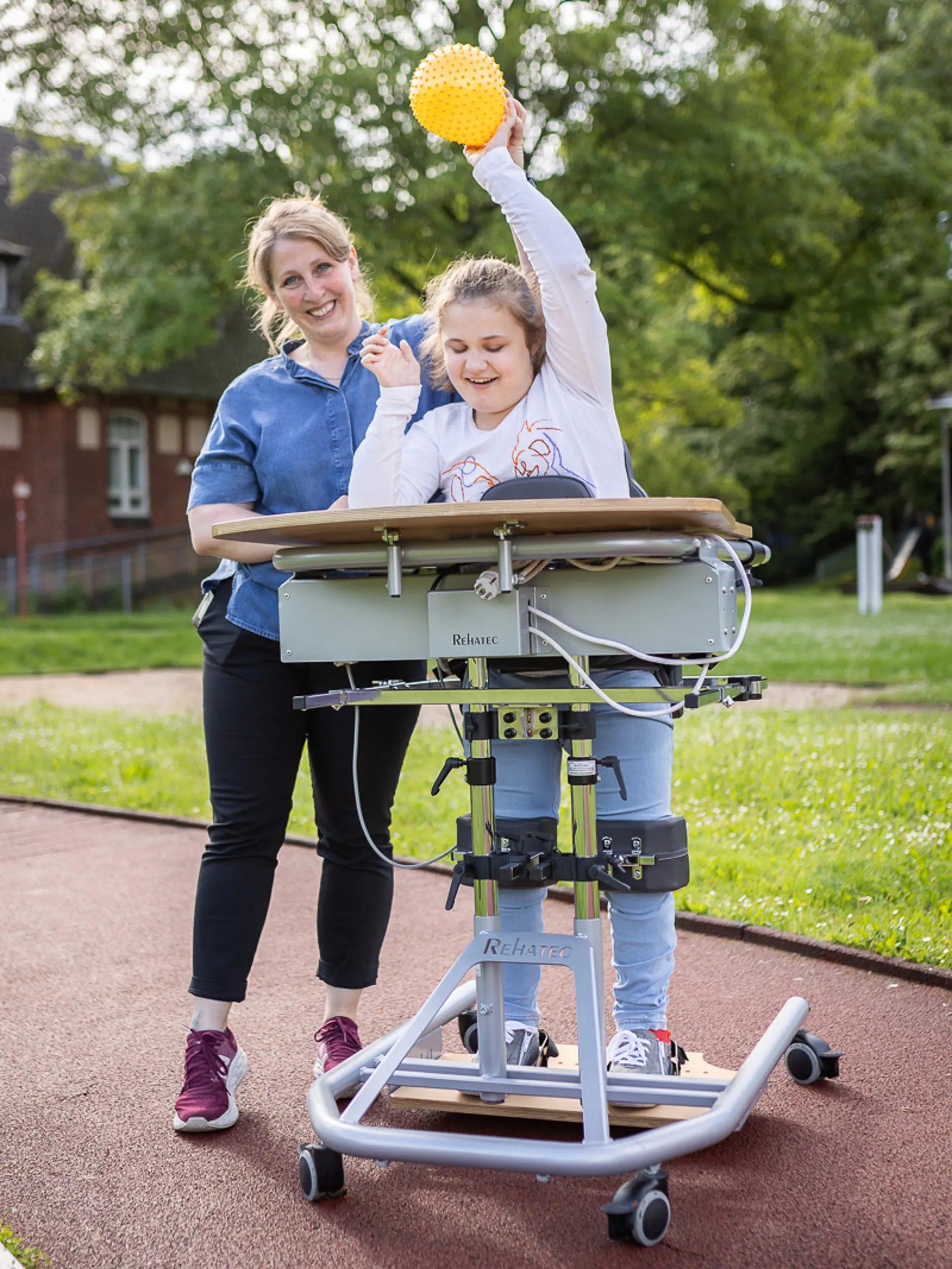 Eine Physiotherapeutin spielt mit einem Kind in einem Stehständer draußen auf dem Schulgelände. Beide lachen und haben Spaß am Spiel.