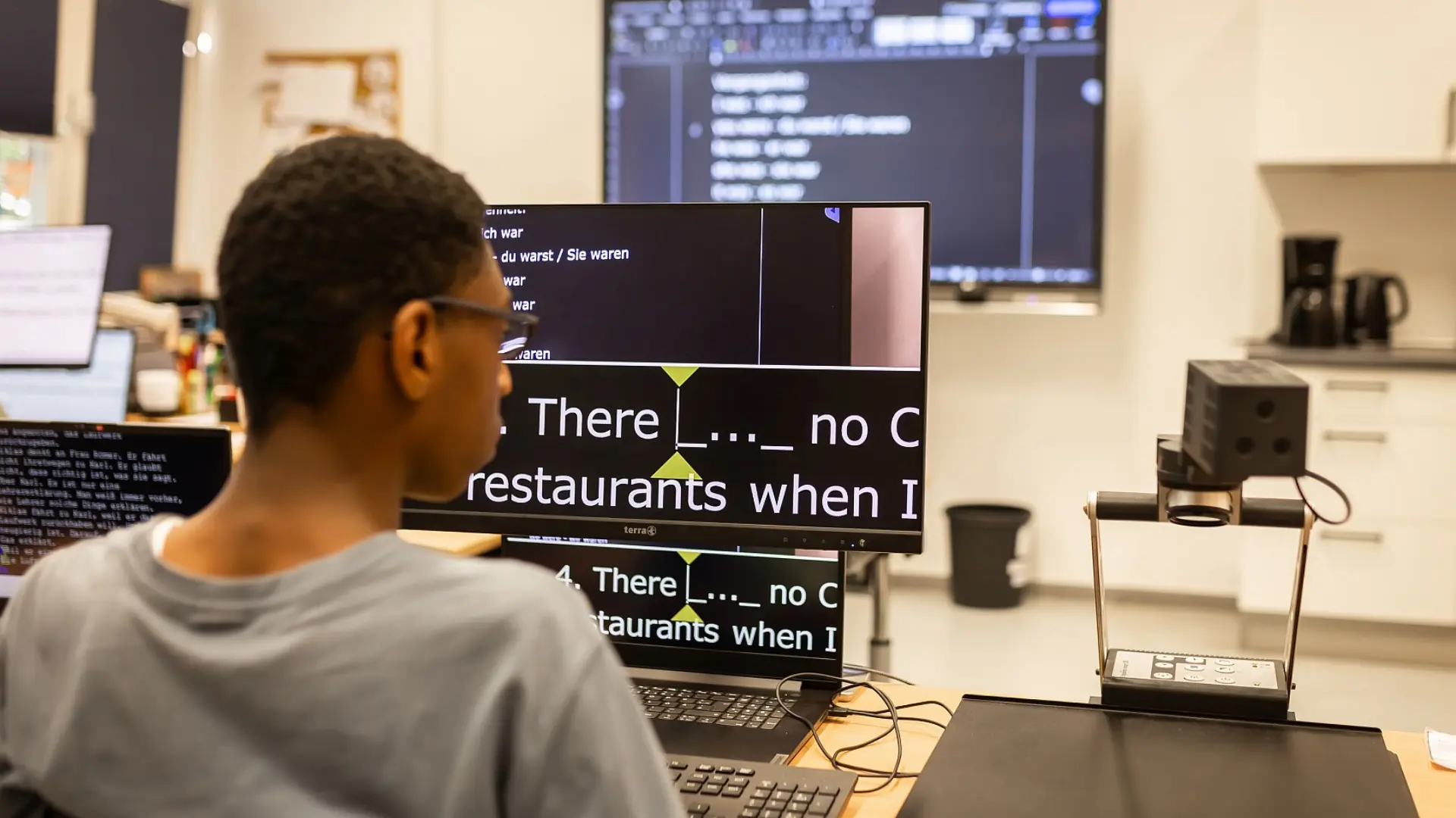 Ein Schüler sitzt am Schreibtisch vorm PC in einem Schulraum. An der digitalen Tafel sind Text zu sehen, der Schüler liest diese an einem speziellen Vergrößerungsdisplay.